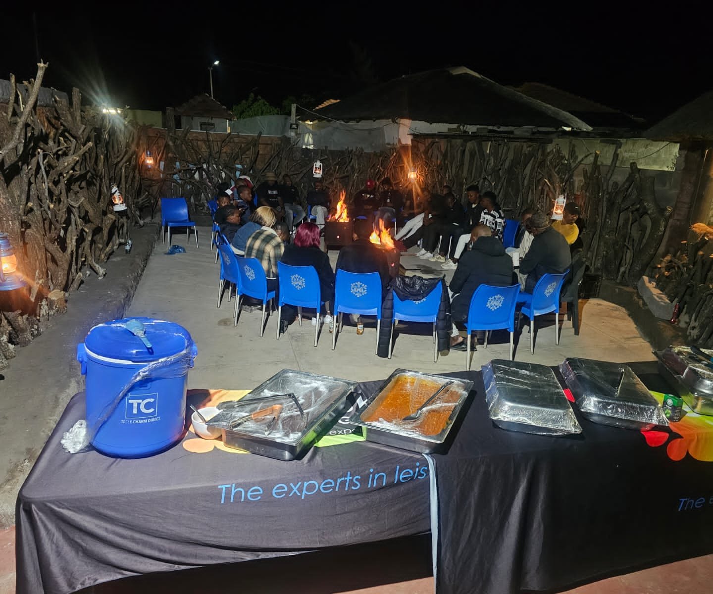 Delegates seated on blue chairs around an open fire in the boma with buffet table in foreground